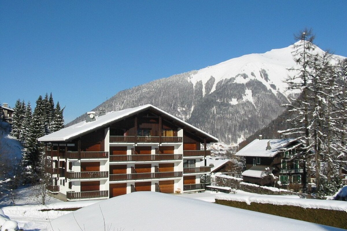 A large wooden chalet with multiple balconies sits in a deep snow-covered mountain village under a clear blue sky, with a snow path.