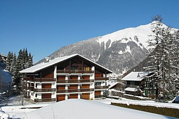 A large wooden chalet with multiple balconies sits in a deep snow-covered mountain village under a clear blue sky, with a snow path.