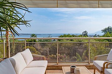 A sunny terrace with white outdoor furniture overlooking a lush garden and the blue sea in the distance.