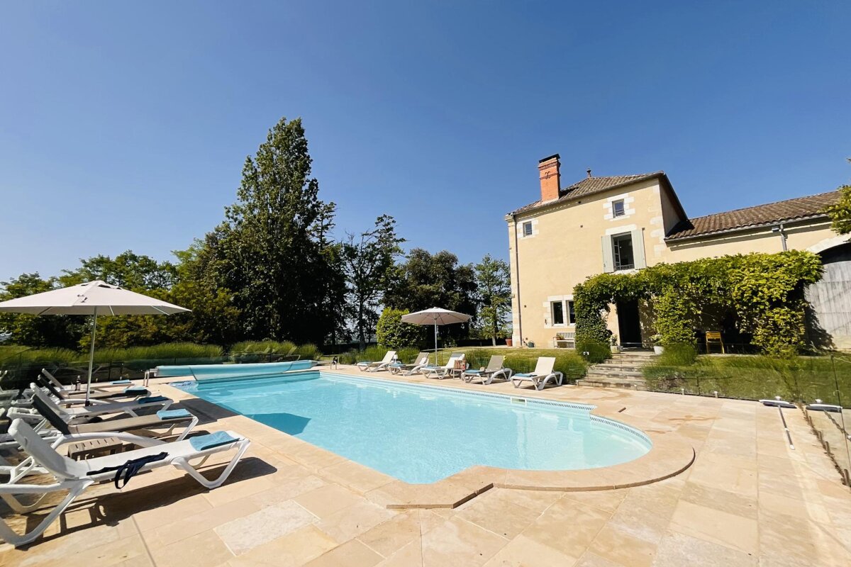 A large outdoor swimming pool with sun loungers and umbrellas on a tiled patio, next to a yellow house with green foliage, all under a bright blue sky.