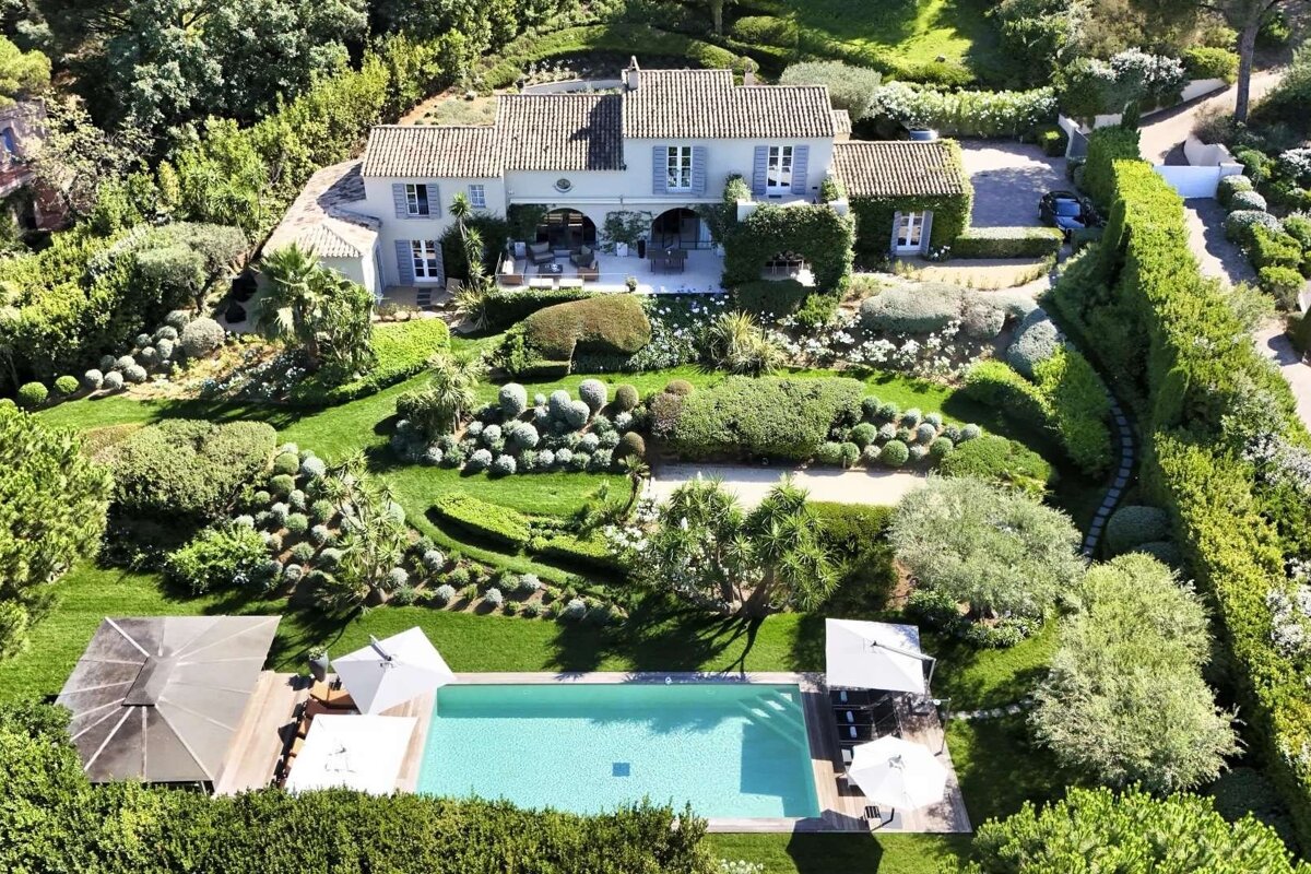 Aerial view of a large, pale villa with an expansive, lush garden featuring manicured topiary and a bright blue swimming pool.