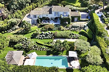 Aerial view of a large, pale villa with an expansive, lush garden featuring manicured topiary and a bright blue swimming pool.