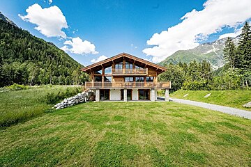 A stunning wooden chalet in a mountain valley, surrounded by lush green grass and dense forests, under a bright blue sky with white clouds.