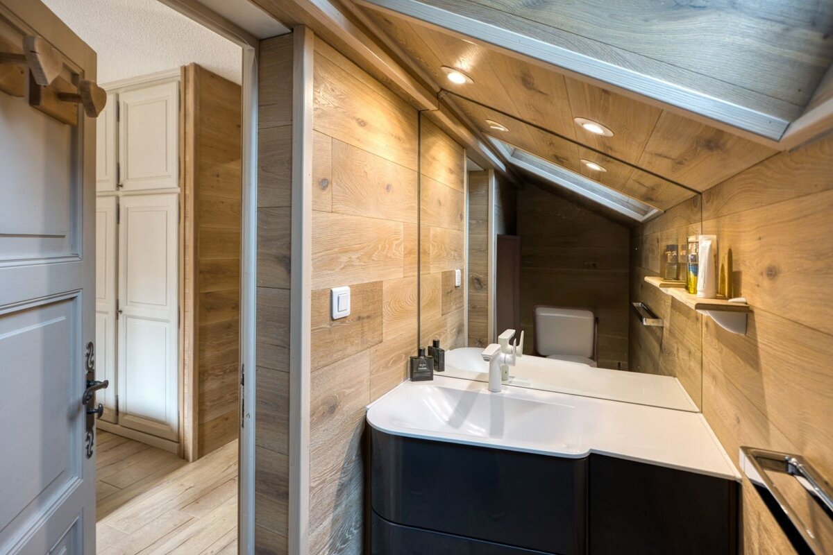 A modern attic bathroom featuring warm wood paneling, a sleek white sink, large mirror, and slanted ceiling. A doorway opens to a hallway.
