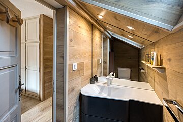 A modern attic bathroom featuring warm wood paneling, a sleek white sink, large mirror, and slanted ceiling. A doorway opens to a hallway.