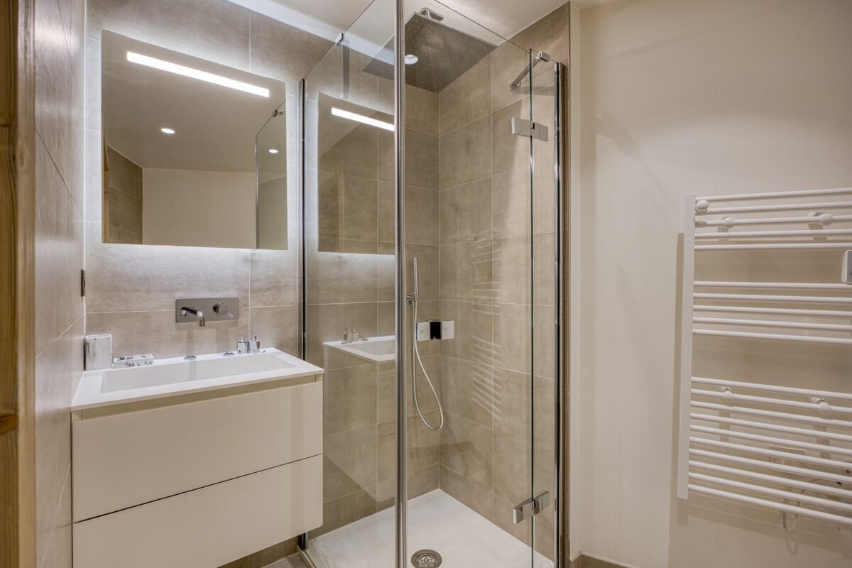 Modern bathroom with a white vanity, lighted mirror, glass shower, and white towel warmer. Neutral tiles and clean design.