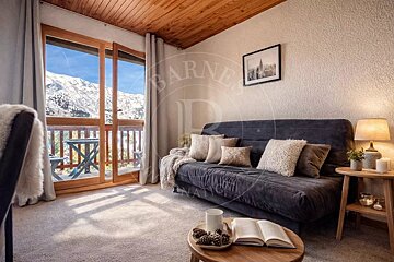 A cozy alpine room with a dark grey sofa, wooden ceiling, and sliding glass doors opening to a snowy mountain view.
