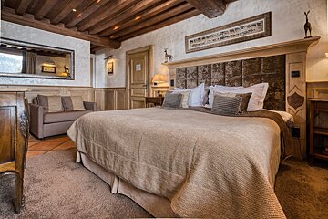 An inviting, rustic bedroom with a large bed, wood-beamed ceiling, plush sofa, and warm earthy tones creating a cozy, lodge-like ambiance.