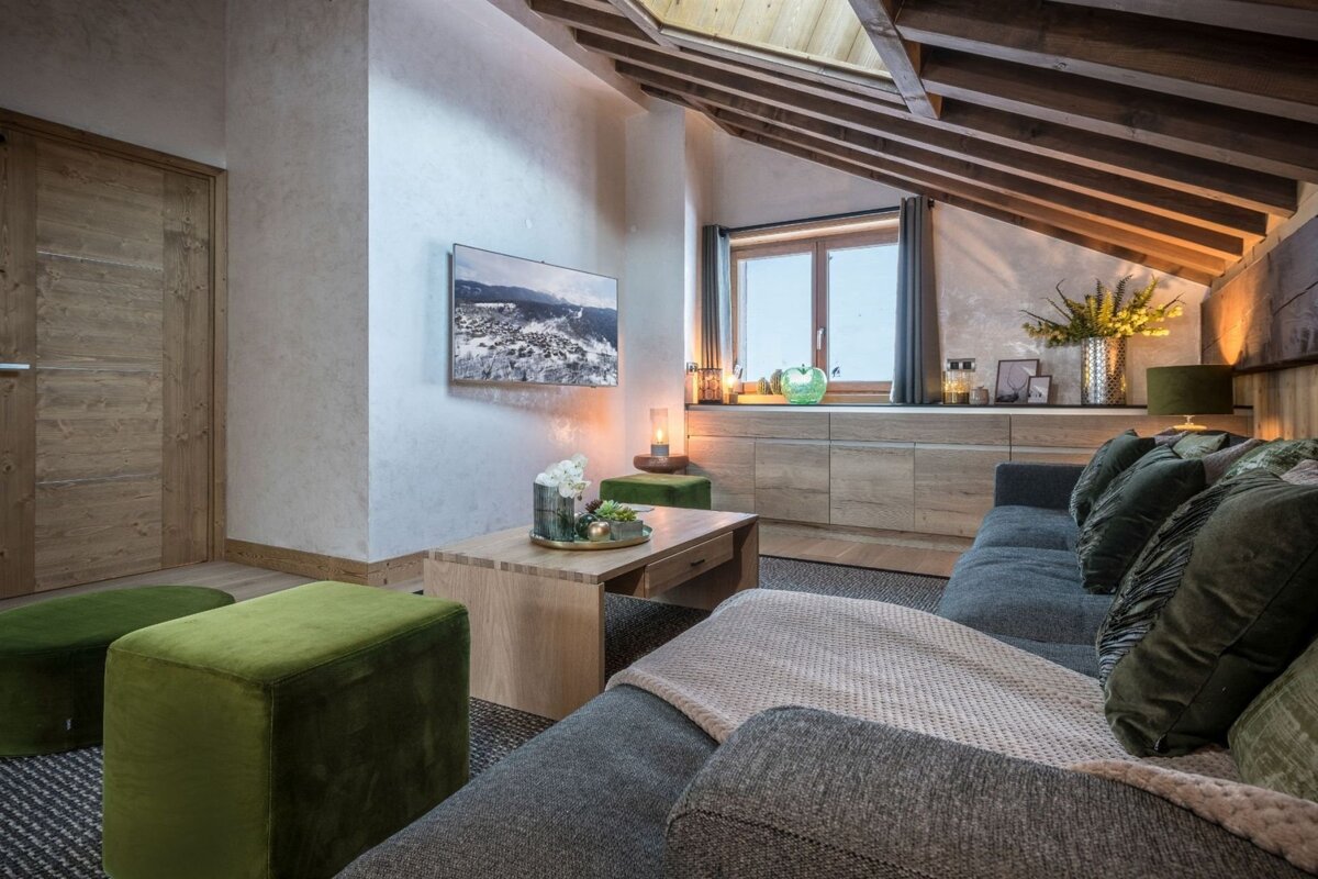 A cozy, modern attic living room featuring a grey sofa, wooden furniture, green accents, a TV, and a window, all under an exposed beam ceiling.