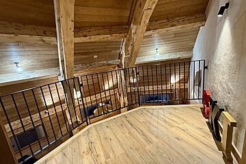 A rustic wooden chalet loft with visible beams, a black metal railing, light wood flooring, and a decorative 
