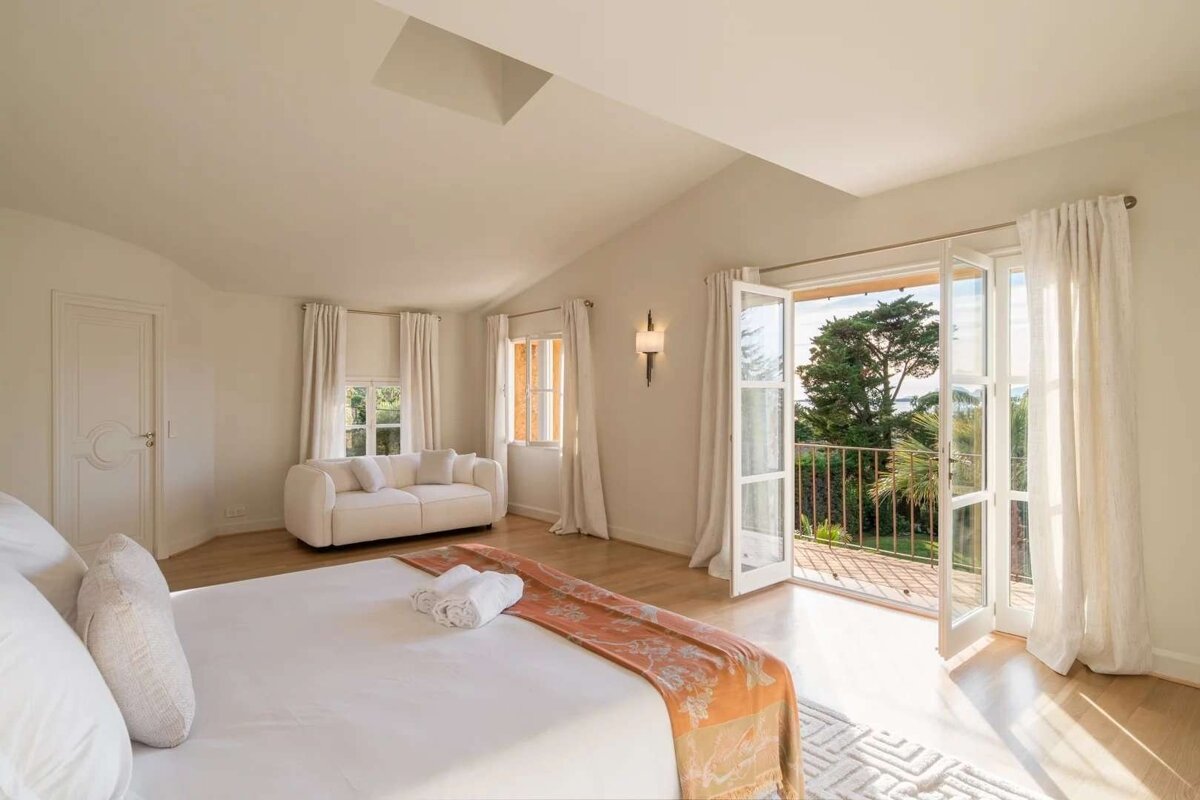 Bright, elegant bedroom with a large bed, white sofa, hardwood floors, and French doors opening to a balcony with a lush green and ocean view.
