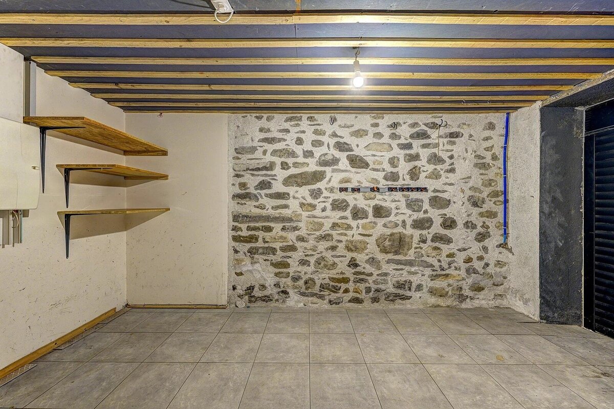 An empty room featuring a prominent stone wall, light grey tiled floor, wooden shelves, an exposed beam ceiling, and a hanging lightbulb.