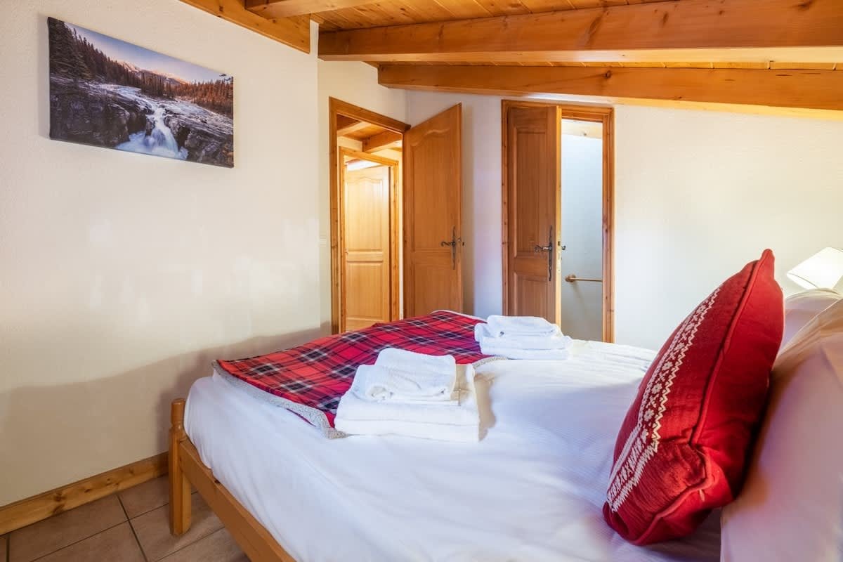 Cozy cabin bedroom featuring a bed with a red plaid blanket, a red pillow, and white towels under a wooden beam ceiling.
