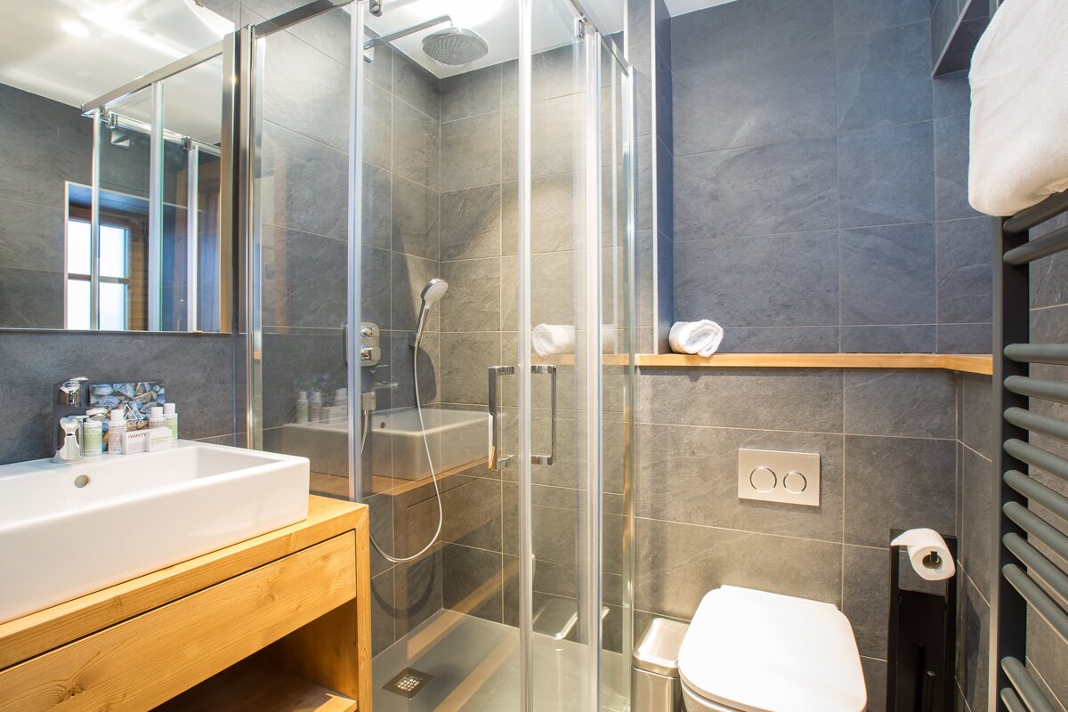 A modern bathroom with dark gray tiles, a wooden vanity and white sink, a glass shower, toilet, and a heated towel rail.