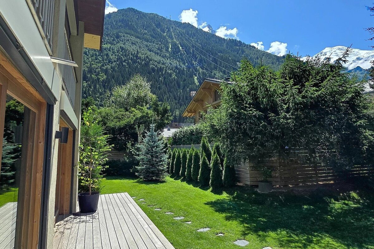 Lush green backyard with a wooden deck and pine trees, set against towering forested mountains and a snow-capped peak.