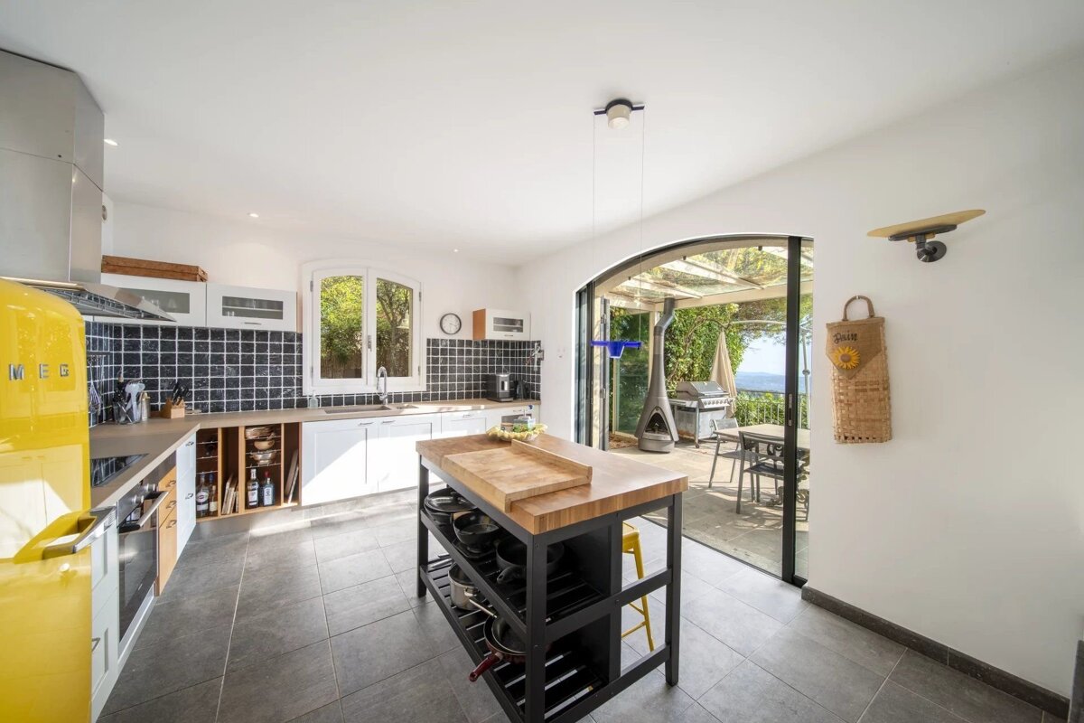 Modern kitchen with a vibrant yellow Smeg fridge, central island, and black tiled backsplash, opening to a patio with a sea view.