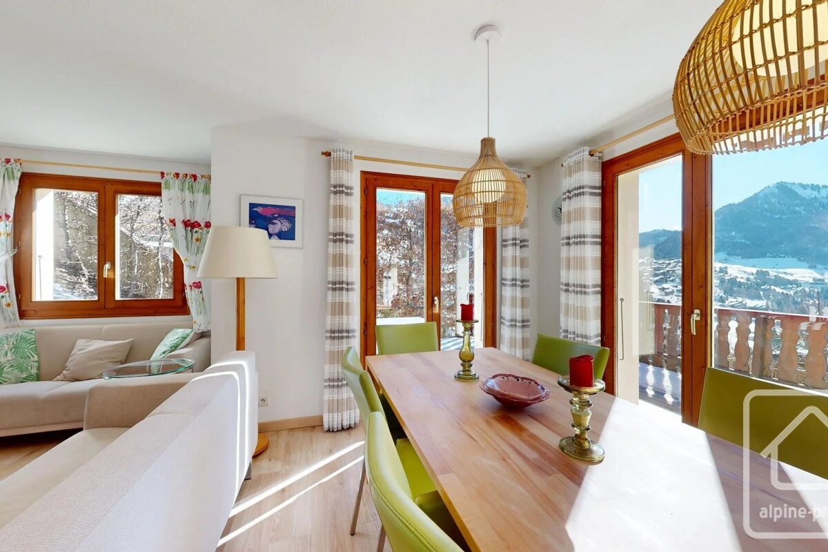 A bright alpine apartment dining area with a wooden table, green chairs, woven lamps, and glass doors opening to snowy mountain views.