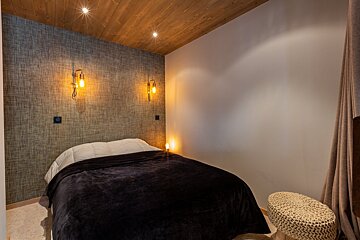 Chalet-style bedroom with a dark bed, rustic textured wall, wooden ceiling, and warm sconce lighting.