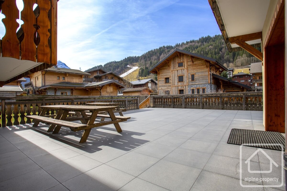 Sunny outdoor patio with picnic tables overlooks an alpine village of wooden chalets and a distant ski slope on a mountain under a blue sky.