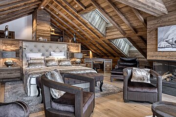 A luxurious, rustic wood-paneled attic bedroom features a large bed with faux fur, comfortable seating, a desk, and a fireplace, illuminated by skylights.
