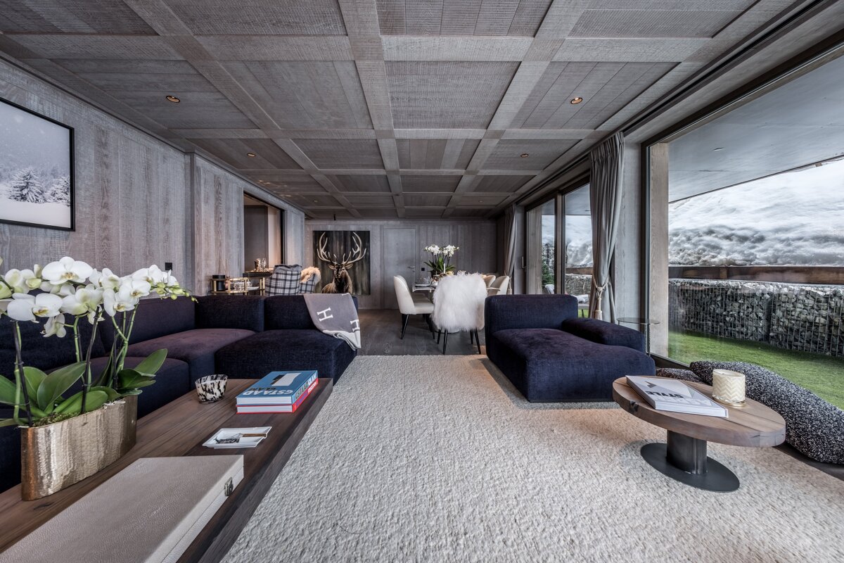 Chic chalet living/dining room with gray wood paneling, dark blue sofas, white orchids, and a large window revealing a snowy mountain vista.