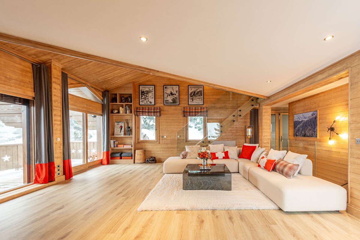 Cozy, modern chalet living room with wood-paneled walls, large cream sectional sofa, red accents, glass staircase, and big windows overlooking snow.