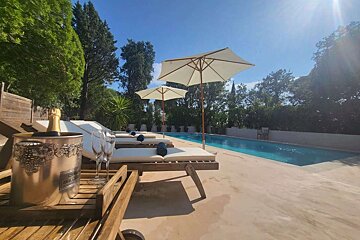 A sunny poolside scene with loungers, umbrellas, and a bucket of champagne with flutes, surrounded by green trees.
