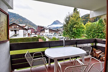Balcony with white table/chairs overlooking a picturesque mountain village, green valley, and misty peaks under a cloudy sky.