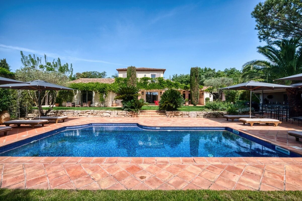 A beautiful villa with a large blue swimming pool, surrounded by terracotta tiles, green garden, loungers, and umbrellas under a bright blue sky.