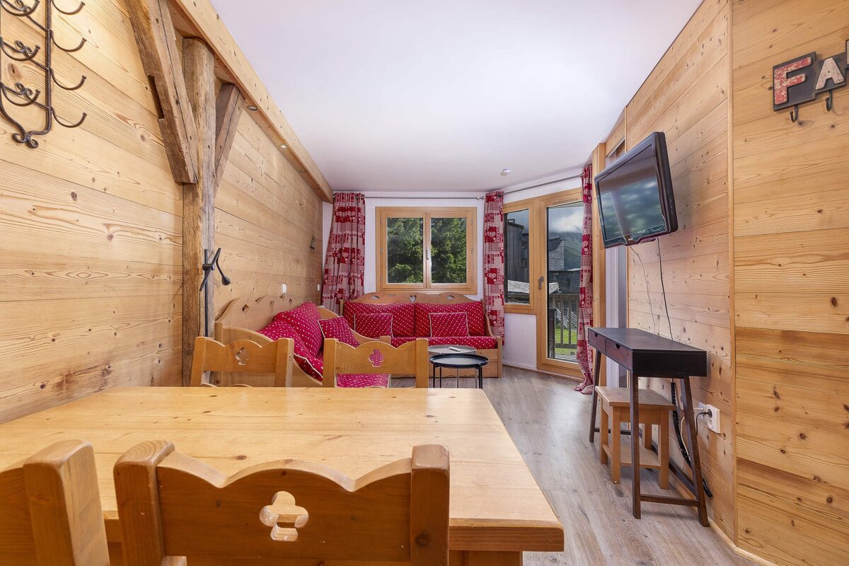 Rustic chalet living room with wood-paneled walls, a large wooden dining table, and red patterned sofas. A window offers a mountain village view.