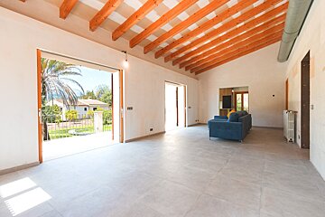 A bright, spacious room with exposed wooden beams and tiled floor. Large doors open to a sunny garden view with palm trees and distant houses. A blue sofa is present.