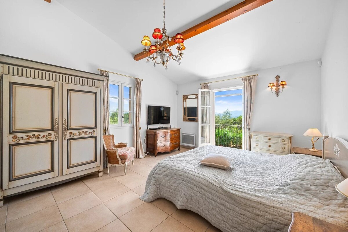 Spacious, bright bedroom with ornate white and wooden furniture, a large bed, and a balcony revealing a lush green view.