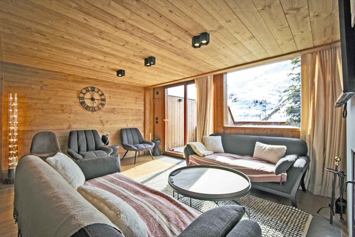 Cozy wooden chalet living room with grey sofas, a round coffee table, and a window overlooking snowy mountain scenery.