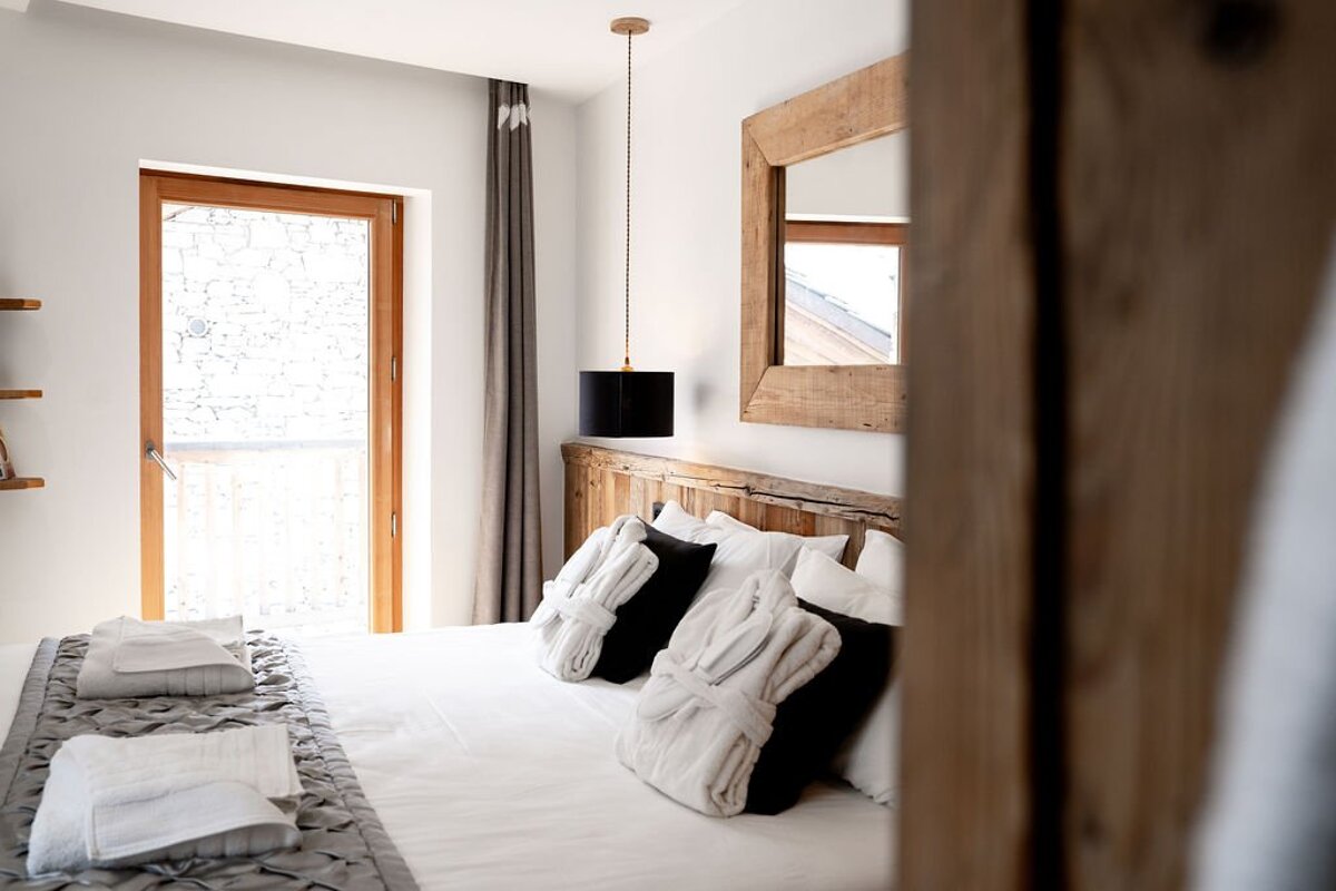 A bright, modern bedroom with white bedding, a wooden headboard, and two white robes. A glass door opens to a sunlit balcony.