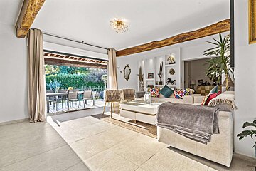 Bright living room with white walls, exposed beams, a light sofa with colorful cushions, and a large opening to a sunny outdoor dining area.