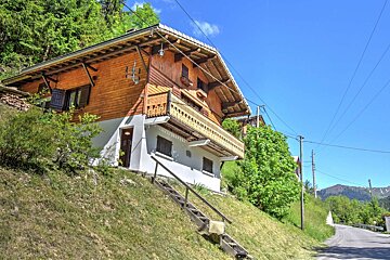 A wooden chalet with a balcony on a steep, grassy hill, surrounded by green trees, under a bright blue sky. A road is visible to the right.