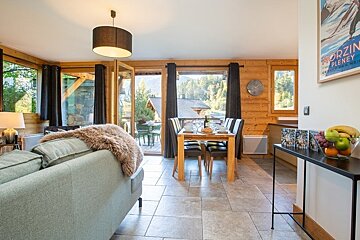 Modern chalet interior featuring a cozy living area with a sofa, a dining set, wooden walls, and large windows with scenic mountain views.