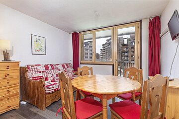 A cozy, chalet-style room with a wooden dining table, bench with red cushions, red curtains, and TV. A large window offers a view of modern apartment buildings.