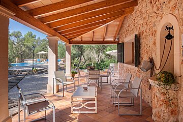 A shaded, rustic stone patio with wooden beams and seating, leading to a sunny pool area nestled among trees, offering a tranquil outdoor retreat.