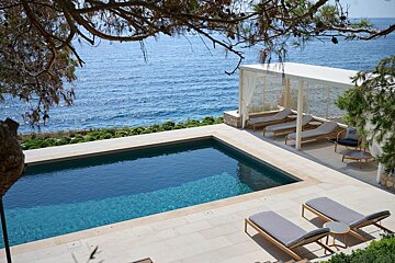 A luxury infinity pool and lounge area overlooking a calm blue sea, framed by pine branches.