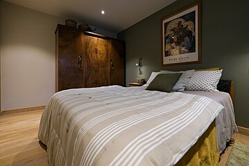 A cozy bedroom featuring a large wooden wardrobe, a bed with a striped comforter, and artwork on a dark green accent wall.