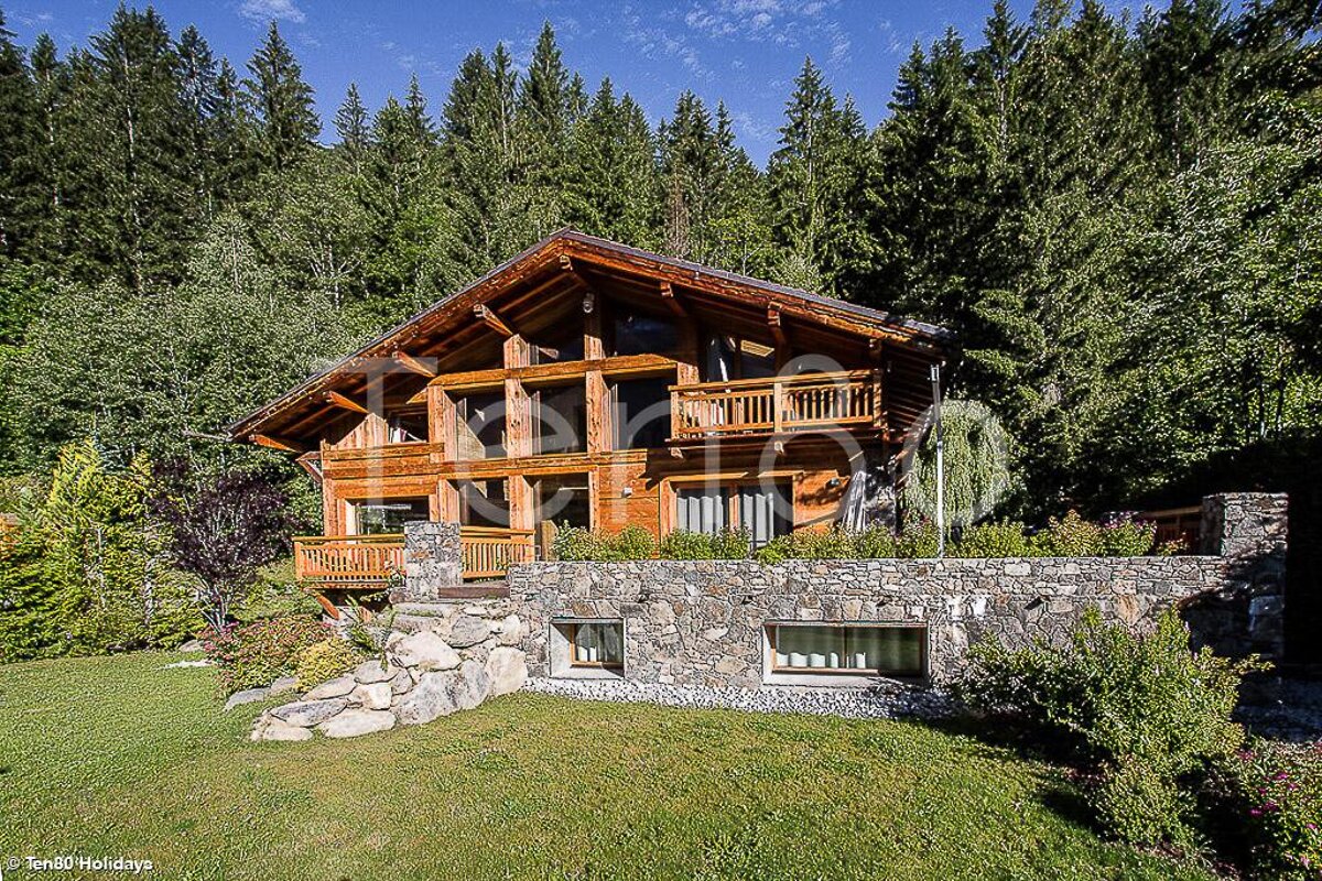 A large wooden house is surrounded by trees and a stone wall