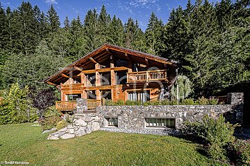 A large wooden house is surrounded by trees and a stone wall