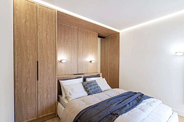 A modern bedroom with light wood built-in cabinetry, a bed with neutral bedding and a blue throw, illuminated by strip lighting.