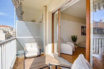 A sunny balcony with a wooden deck, outdoor seating, and a view of buildings, connected to a modern indoor living space via a sliding glass door.