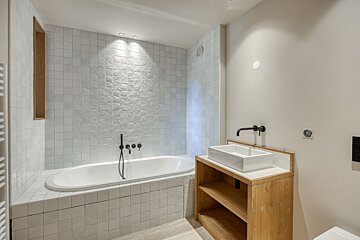 Modern bathroom with a tiled built-in tub, black hardware, and a wooden open-shelf vanity with a white sink.