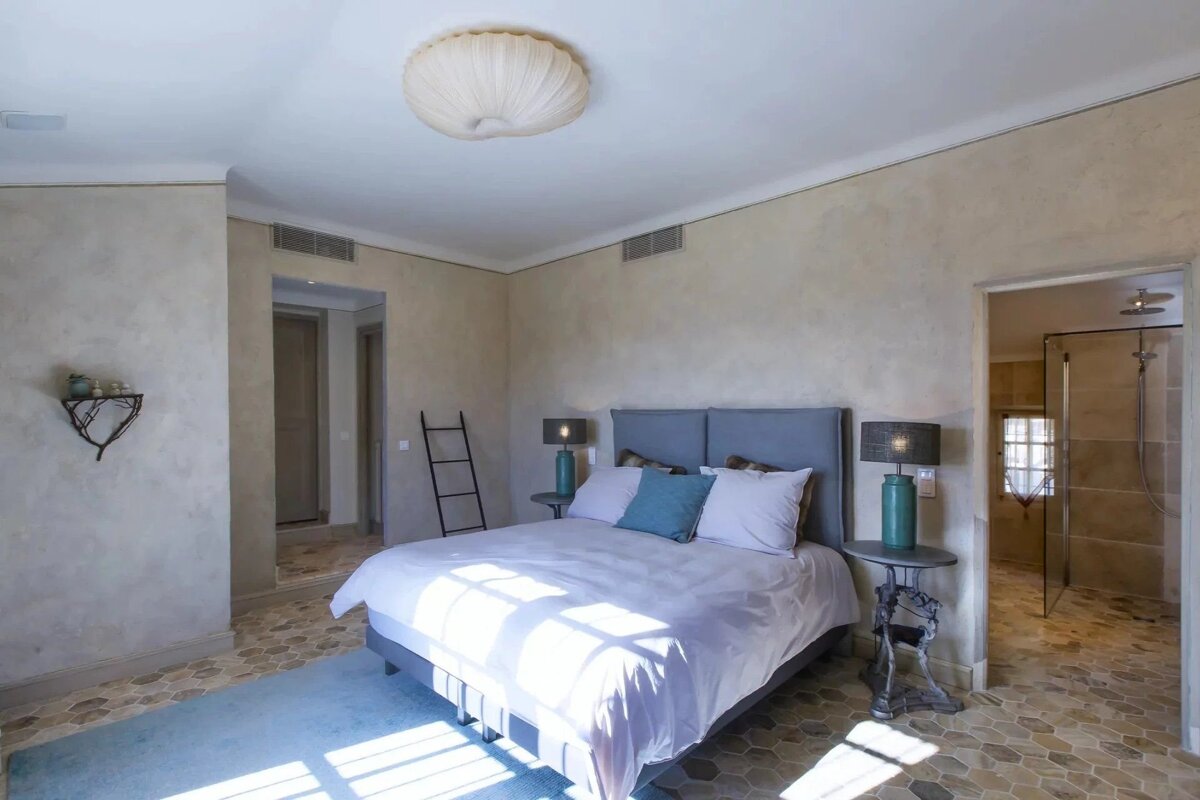 Sunlit bedroom with a grey bed, hexagonal tiled floor, and an open doorway leading to a bathroom with a glass shower.