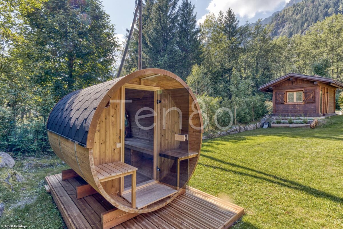 A wooden barrel sauna on a grassy lawn, with a traditional wooden cabin and dense forest in the background under a bright sky.