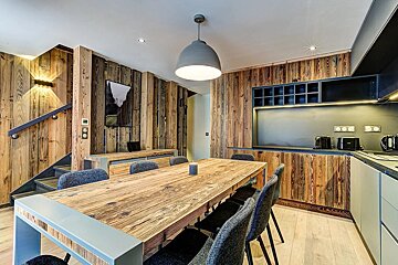 A rustic modern dining and kitchen area featuring wood-paneled walls, a large wooden table with grey chairs, a sleek kitchen, and a staircase.