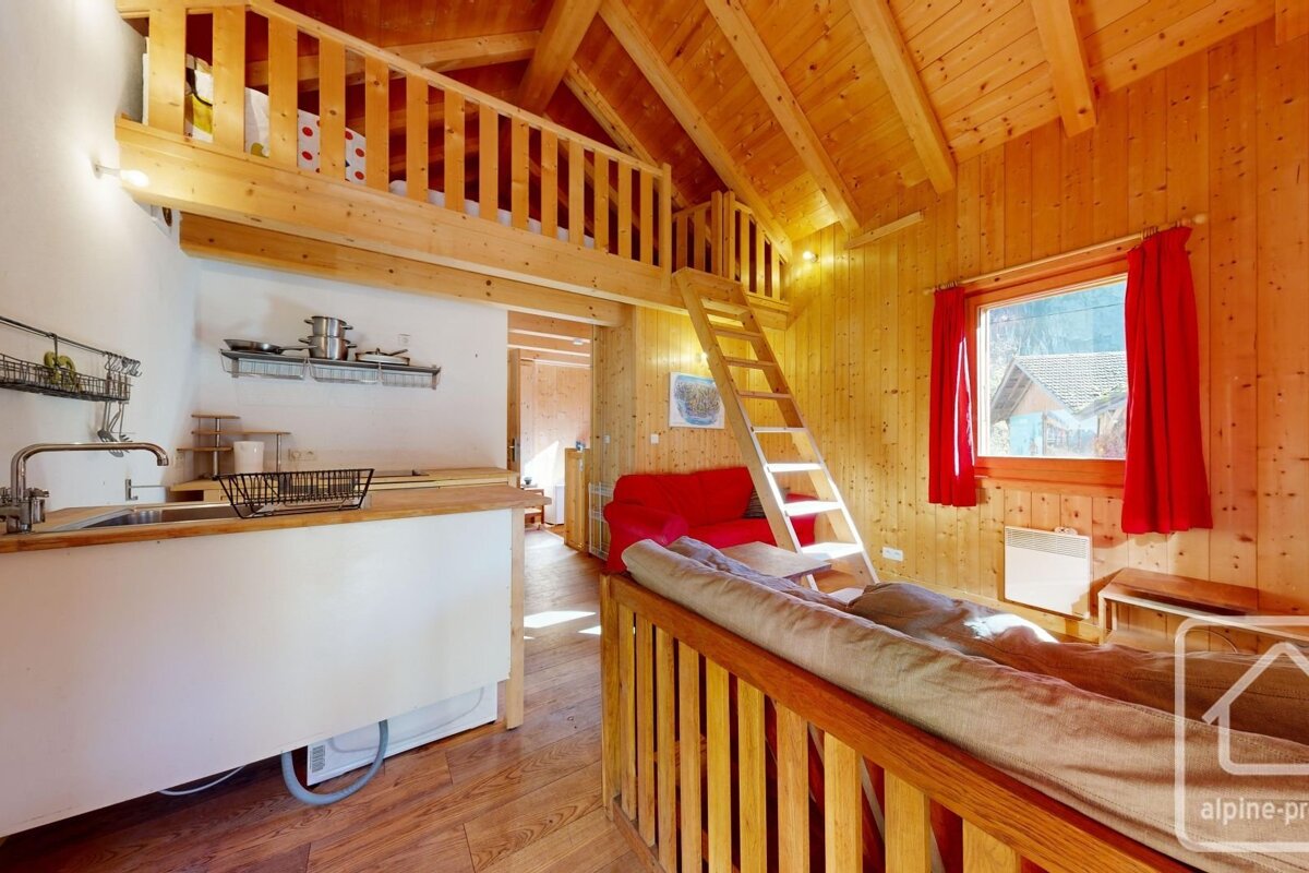 A cozy, rustic wooden cabin interior with a loft accessible by a ladder, a compact kitchenette, and a living area featuring a red sofa and red-curtained window.